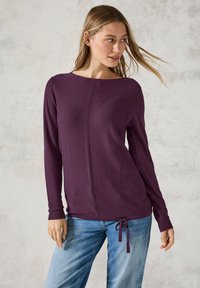 Lila Pullover mit langen Ärmeln und geripptem Muster, runden Ausschnitt und verstellbarem Bindeband am Saum, kombiniert mit hellblauen Jeans.