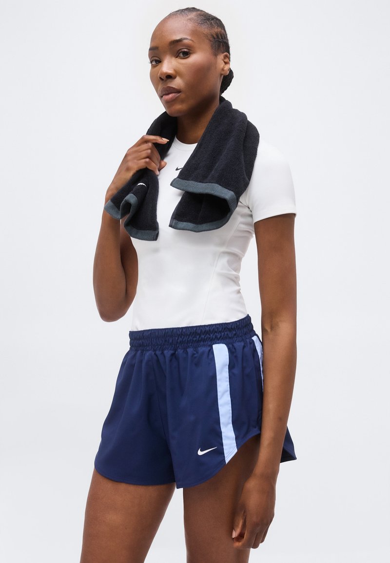 Mujer atlética sosteniendo una toalla negra alrededor del cuello, vistiendo una camiseta blanca ajustada y pantalones cortos azul marino de Nike con rayas azul claro.