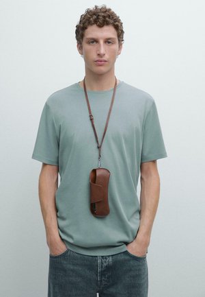 Maglietta in cotone verde chiaro a maniche corte, abbinata a jeans scuri in denim. Un portadocumenti in pelle marrone pende da una tracolla intorno al collo.