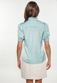 Blusa di raso azzurro chiaro con maniche a sbuffo e colletto arricciato, indossata con una gonna cremosa testurizzata con sottili decorazioni.
