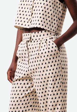 Persoon draagt witte hoge taille broek en bijpassende mouwloze cropped top met zwart onregelmatig stippenpatroon, één hand in de zak.