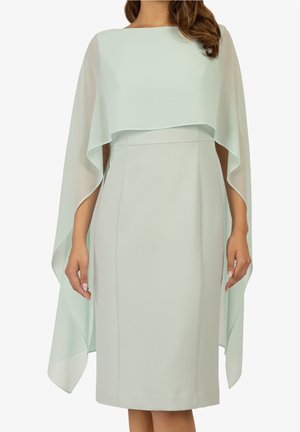 Vrouw draagt een lichtgroene midi-jurk met doorschijnende cape-overlay en mouwen van middelbare lengte, staand tegen een effen achtergrond.