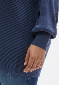 Μπλε σκούρο ριπ πουλόβερ με μακριά, φαρδιά μανίκια και υφή, με κοντινή λήψη ενός χεριού που ακουμπά στη ραφή του πλαινού.