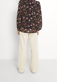Blusa floreale nera con motivi colorati, maniche lunghe e polsini elasticizzati; abbinata a pantaloni in velluto a coste chiari a gamba larga.