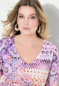 Lila gemusterte V-Ausschnitt-Bluse mit lebhaften Designs, verschiedenen Formen und subtilen Akzenten. Das Model trägt silberne Creolen.