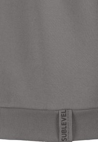 Grauer Hoodie mit weichem Stofftextur, geripptem Saum und einem seitlichen Etikett mit "SUBLEVEL"-Markierung. Einfaches, funktionales Design.