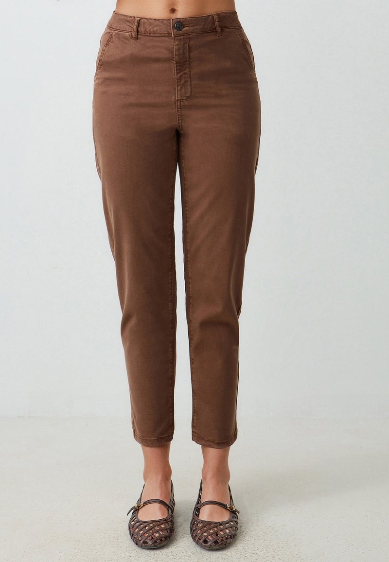 Pantaloni in cotone marrone con una vestibilità slim, tasche frontali e chiusura con bottoni, che terminano sopra la caviglia. Abbinati a scarpe ballerine intrecciate marroni.