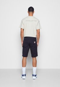 T-shirt bege de mangas curtas com texto nas costas, combinado com calções cargo azul-marinho com bolsos laterais e um logo. Meias brancas e ténis.