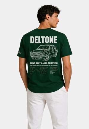 Uomo che indossa una maglietta verde scuro con illustrazione bianca di un'auto e specifiche dettagliate stampate sulla schiena, abbinata a pantaloni bianchi.