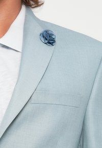 Blazer bleu clair avec une finition texturée, doté d'une poche poitrine simple et d'une épingle en tissu bleu en forme de fleur sur le revers.