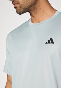 Svetlo modra športna majica iz teksturirane tkanine, z črnim trikotnim logotipom na zgornjem levem delu prsnega koša in okroglim vratnim izrezom.