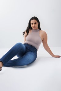 Bodysuit de punto beige con cuello alto, combinado con jeans de tiro alto en azul oscuro. Modelo sentada sobre un fondo blanco, luciendo tacones negros.