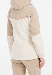 Veste à capuche bicolore beige et blanc cassé portée avec un pantalon beige, vue de dos avec poignets élastiques et poche zippée sur le pantalon.