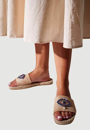 Füße, die beige geflochtene Sandalen mit blau-rosa verzierten Augenmotiven tragen, kombiniert mit einem hellbeigen Rock.