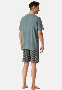 T-shirt à manches courtes teal clair associé à un short à carreaux dans des couleurs vert, marron et crème. La personne se tient pieds nus, de dos.