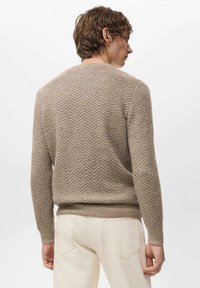 Maglione beige lavorato a maglia con motivo a spina di pesce, scollo rotondo e polsini e orlo a costine, indossato con pantaloni di colore chiaro.