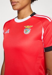 Rød sportsdrakt laget av teksturert stoff med svart og hvite detaljer; har Benfica-logoen på venstre bryst og Adidas-merke.