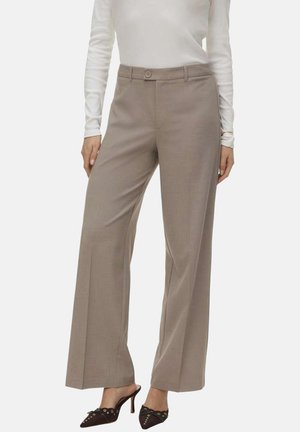 Femme portant un pantalon taupe taille haute à jambes larges, associé à un haut blanc à manches longues et des chaussures à talons pointues foncées.