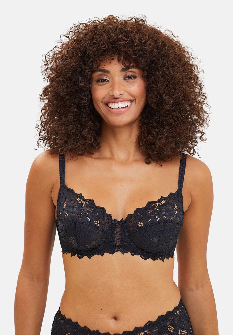 Reggiseno in pizzo nero con bordi ondulati, motivi floreali e un centro in rete. Presenta spalline regolabili e chiusura a gancio e occhiello sul retro.