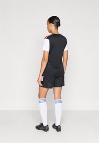 Μαύρο αθλητικό μπλουζάκι με κόκκινες και λευκές λεπτομέρειες, συνδυασμένο με μαύρο σορτς, λευκές κάλτσες με μπλε ρίγες και μαύρα ποδοσφαιρικά παπούτσια.