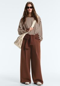 Frau trägt ein braun-beige gestreiftes Langarmshirt, weite braune Hose, Sonnenbrille, weiße Turnschuhe und eine cremefarbene Schultertasche.