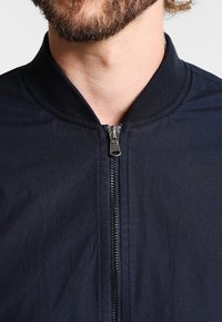 Marinblå bombarjacka med dragkedja, ribbad krage och slät yta, med minimalisk design och diskreta detaljer.