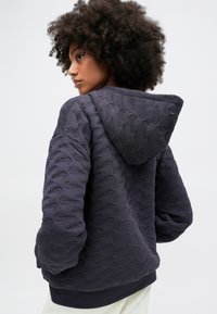 Ciemnofioletowa pikowana bluza z kapturem o teksturowanym wzorze, luźny krój i kaptur. Posiada subtelny detal z logo na plecach w pobliżu kołnierza.