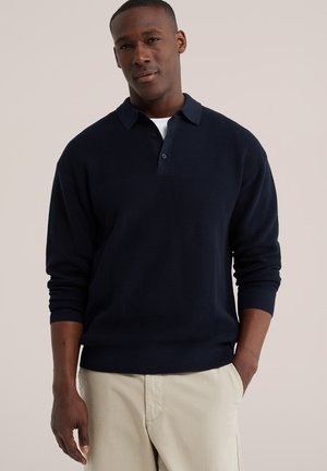 Homme portant un pull polo bleu marine à manches longues et un pantalon beige, debout avec une main dans la poche, sur un fond neutre.