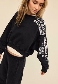 Felpa nera corta con testo bianco sulla manica. Materiale morbido, vestibilità ampia e polsini a costine. Abbinata a pantaloni neri coordinati.