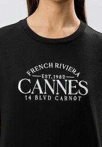 Musta puuvillapaita, jossa on valkoista brodeerattua tekstiä: "FRENCH RIVIERA CANNES EST. 1982 14 BLVD CARNOT" rinta-alueella. Pyöreä kaula-aukko.