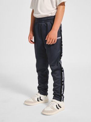 Kind draagt zwarte Hummel trainingsbroek met witte logostrepen en witte sneakers, staand op een effen achtergrond.