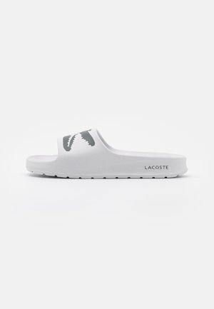 Biała sandałka Lacoste typu slide z ciemnozielonym logo krokodyla na pasku oraz oznaczeniem Lacoste po zewnętrznej stronie podeszwy.