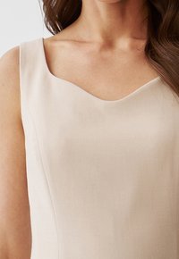 Beige ärmlös klänning med rundad halsringning, med en texturerad tyg och en figurnära silhuett som visar mjuka konturer.