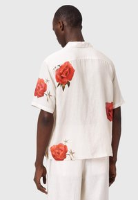 Hombre con camisa blanca de manga corta y pantalones con grandes estampados de rosas rojas en la espalda y las mangas, de pie frente a un fondo liso.