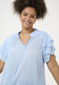 Light blue blouse featuring ruffled sleeves, a V-neck with a keyhole detail, and a smooth, lightweight fabric. Soft texture and relaxed fit.