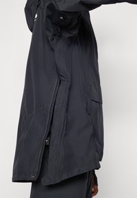 Abrigo rompevientos largo de color gris oscuro con una textura suave, que presenta bolsillos laterales, un cierre de cremallera y un ligero brillo.