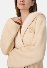 Beżowy szlafrok z miękką, teksturowaną podszewką wewnętrzną. Posiada kołnierz szalowy oraz haftowane logo DKNY. Długie rękawy z szerokimi mankietami.