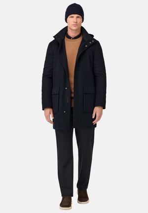 Parka in misto lana blu navale con colletto alto, due tasche frontali e dettagli con bottoni; maglione marrone e pantaloni scuri; scarpe slip-on marroni.