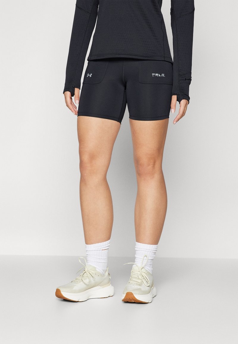 Personne portant un short de sport noir, un haut à manches longues noir, des chaussettes blanches et des chaussures de course beige clair, debout sur un fond clair uni.