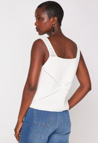 Femme aux cheveux courts portant un haut sans manches en maille blanche et un jeans bleu, de profil avec la main sur la hanche contre un fond uni.