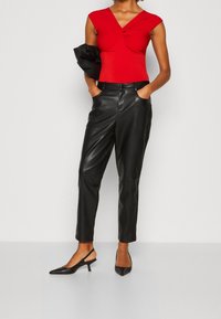 Kobieta ubrana w dopasowaną, czerwoną bluzkę z krótkimi rękawami, czarne skórzane spodnie z wysokim stanem, czarne sandały na obcasie, trzymająca czarną kurtkę.