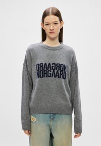 Suéter cinza de malha com decote redondo, ombros caídos e o logotipo "Norgaard" em azul escuro na parte da frente. Usado com jeans azul desbotado.