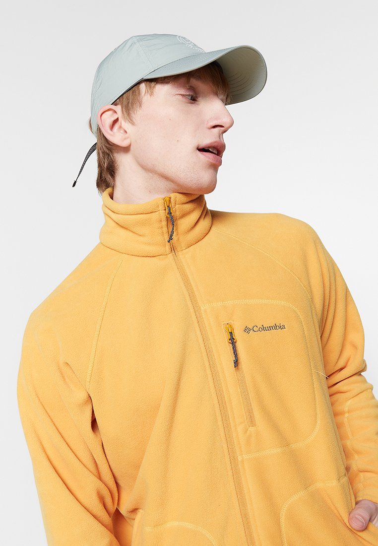 Gelbe Fleecejacke mit einer Reißverschlusstasche, hohem Kragen und gelbem Reißverschlussz pull. Die Person trägt eine hellgraue Mütze.