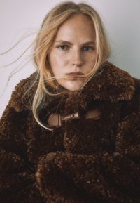 Hnedý kožušinový kabát s plyšovou textúrou, veľkým golierom a koženými detailmi. Dizajn je nadrozmerný s viditeľnými švami.