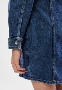 Τζάκετ τζιν με εφαρμοστό σχέδιο, μπλε χρώμα, ορατή ραφή και κουμπί σνάπ στο μ�ανσέτα. Διαθέτει πλαϊνή τσέπη.