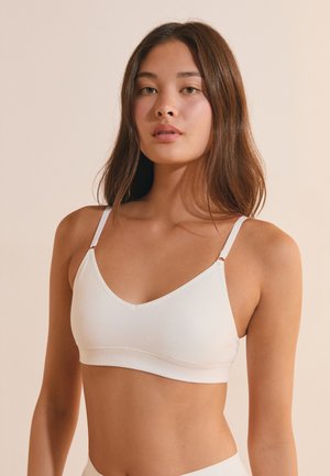 Jeune femme aux longs cheveux bruns portant un bralette blanc sur un fond beige uni, faisant face avec une expression neutre.
