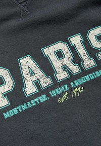 Tmavě šedá látka obsahuje velké, opotřebované bílé nápisy s textem "PARIS" s tyrkysovými a žlutými akcenty, což naznačuje vintage styl.