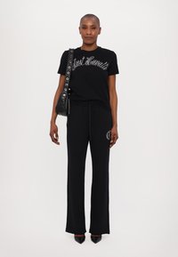 Juodas marškinėliai su metaliniu užrašu, derinami su juodais sportiniais džinsais, kurie turi grafiką. Aksesuarai apima juodą maišą su sidabro detalėmis.