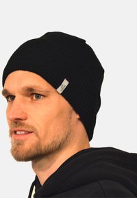 Black ribbed beanie hat made of knitted fabric, featuring a small silver logo tag on the side, and a close-fitting shape.