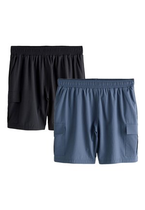 Mężczyzna stojący z rękami za plecami, ubrany w białą koszulkę, czarne szorty, białe skarpety i białe buty sportowe na białym tle.